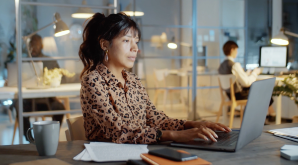 A woman working on a laptop in an office with a glass partition. Papers, a phone, and notebooks are on the desk. Another person is in the background.